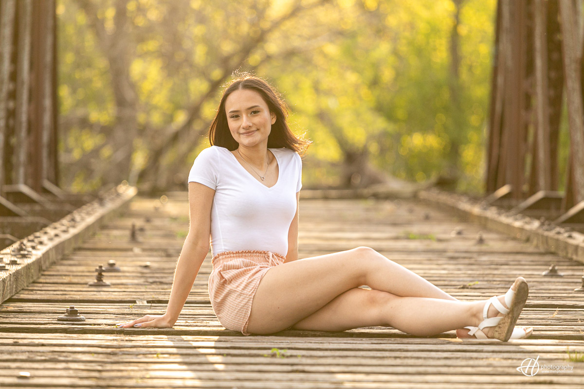 High school senior standing on a old bridge at sunset, looking off into the distance with a smile. The warm light of the sunset creates a golden glow, casting long shadows on the bridge