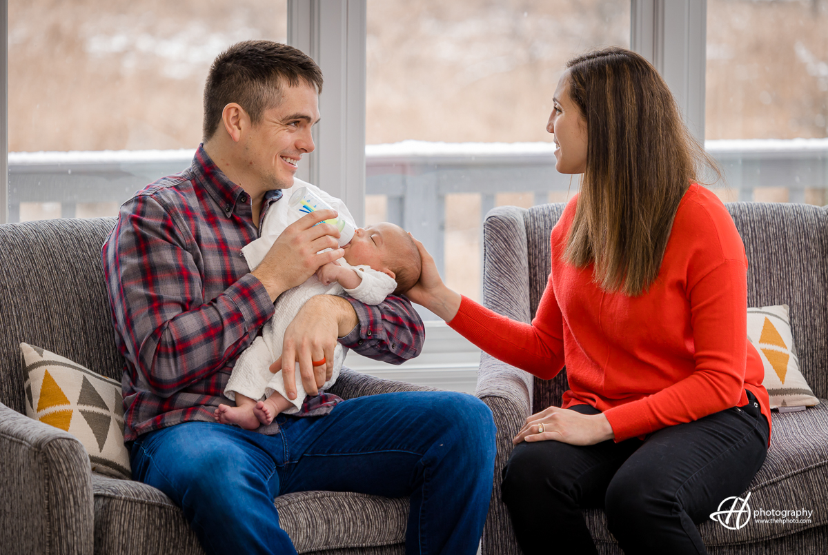 A heartwarming family moment is captured in this photo as a mother and father sit side-by-side, cradling their newborn baby in their arms. The baby is feeding from a bottle, while the parents look down at their little one with love and tenderness.