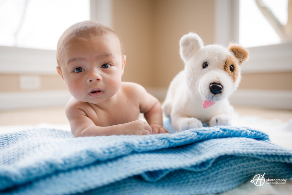 a newborn baby is captured sitting up and looking straight at the camera, with a dog toy visible in the background. The image conveys a sense of curiosity and attentiveness, as well as a feeling of warmth and comfort. The photo is both charming and heartwarming