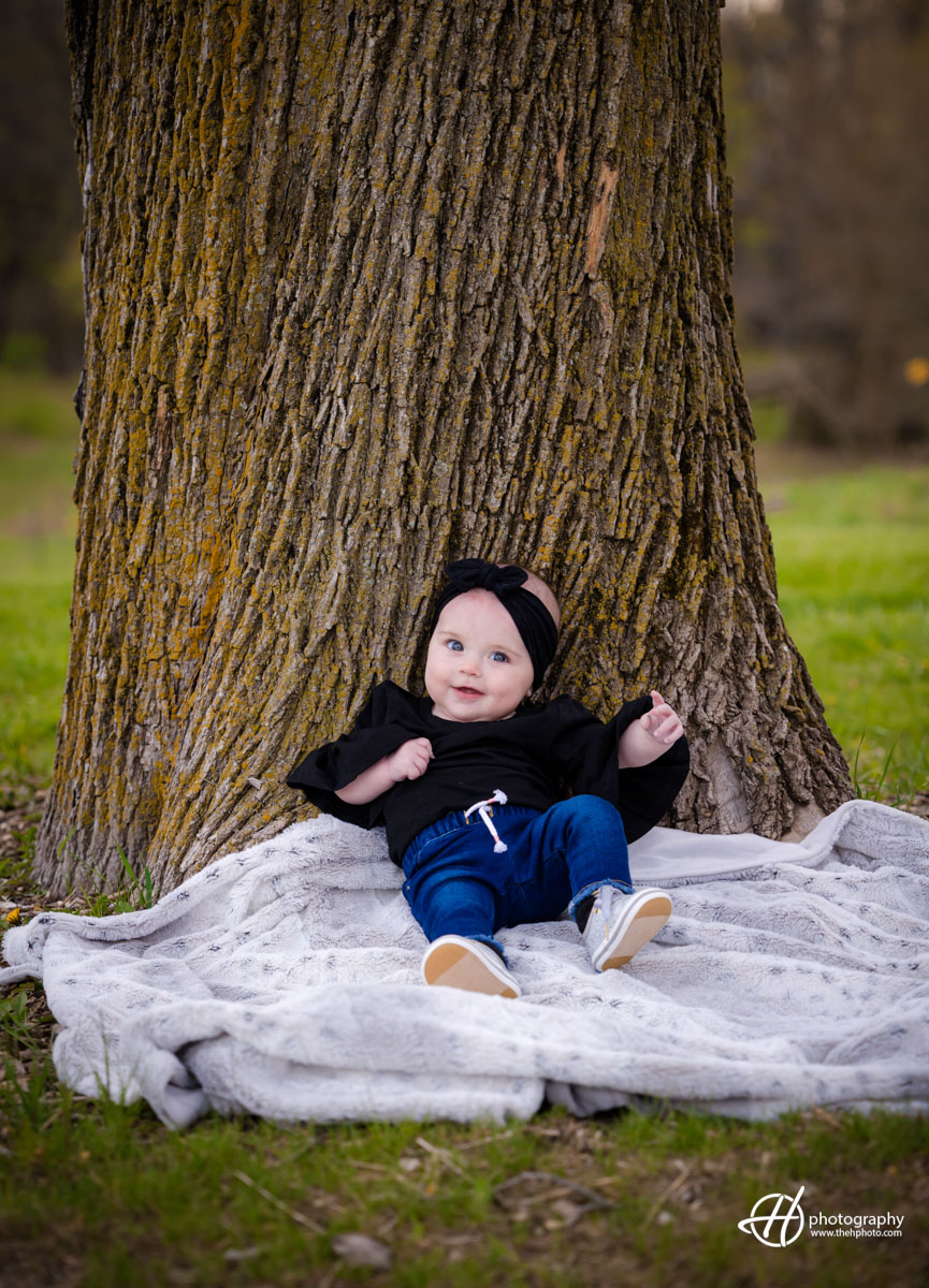 Baby Blake leaning against a sturdy tree trunk in the forest. Baby Blake, dressed in an adorable outfit, leans comfortably against the rough texture of the tree. Her tiny hands grasp the bark, while her curious gaze explores the surrounding natural beauty. Sunlight filters through the branches above, casting a warm glow on her cherubic face. The serene woodland backdrop creates a peaceful atmosphere, capturing a moment of quiet contemplation and wonder in nature.