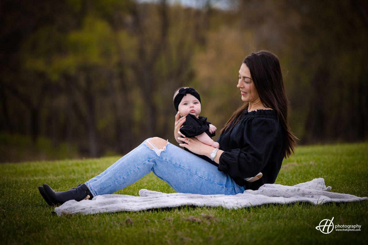 A touching image capturing the bond between a mother and Baby Blake. The mother sits on her knees, her arms cradling Baby Blake gently. Both mother and baby share radiant smiles, radiating love and happiness. The soft green grass serves as a natural carpet beneath them, while colorful spring blossoms add a vibrant touch to the scen