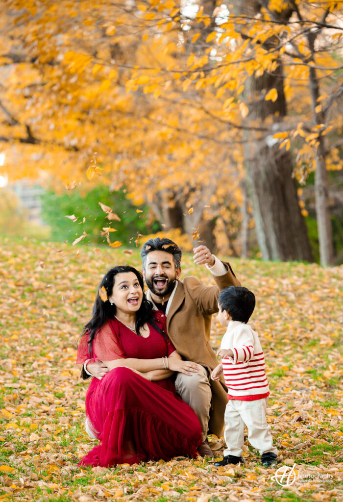 Wholesome family fun as the family joyfully plays amidst falling leaves in the park during their photo session. Reidan's laughter, Nimi's radiant smile, and Roshan's playful engagement create a heartwarming scene against the backdrop of autumn foliage. A delightful capture of familial happiness in motion.