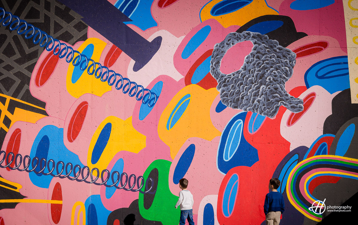 Dorian and Theo captivate the scene against a vibrant wall painting, surrounded by a burst of colors. Their playful spirits come alive as they interact with the artwork, creating a lively and dynamic moment frozen in time. The rich hues of the mural provide a striking contrast, highlighting the joy and energy of this delightful sibling duo.