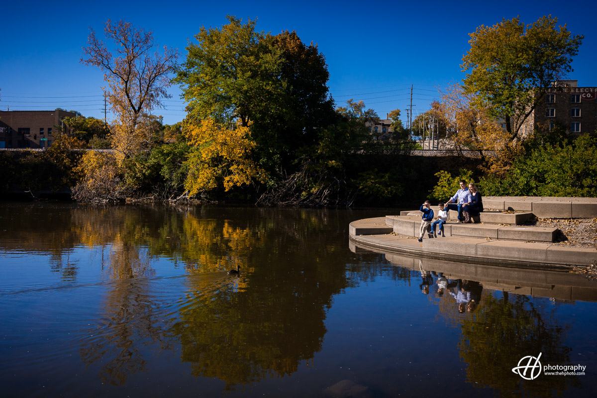 he Woodcox family stands united, gazing fondly at the Fox River from the scenic Walton Island Park in Elgin, IL. Fall foliage frames the backdrop, casting a warm glow on their faces. The connection between family members is palpable against the tranquil river, capturing a moment of shared serenity and appreciation for nature's beauty