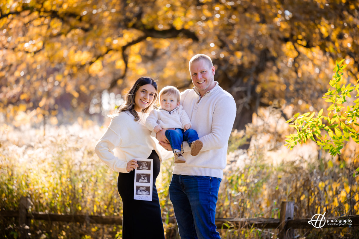 A radiant fall day in Barrington as Susana, Dan, and their firstborn share a heartwarming moment surrounded by golden leaves. The family embraces amidst the enchanting autumn foliage, capturing the joy and anticipation of welcoming a new member into their loving fold. The warm glow of fall sunlight adds a magical touch to this beautiful family scene.