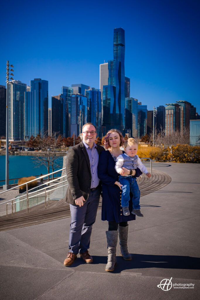Family joy at Navy Pier: Sara, Todd, and their son, adorned in festive and fall attire, share a moment of connection against the breathtaking backdrop of the Chicago skyline. The iconic Navy Pier arch and Lake Michigan provide a picturesque setting, capturing the essence of their annual family tradition.