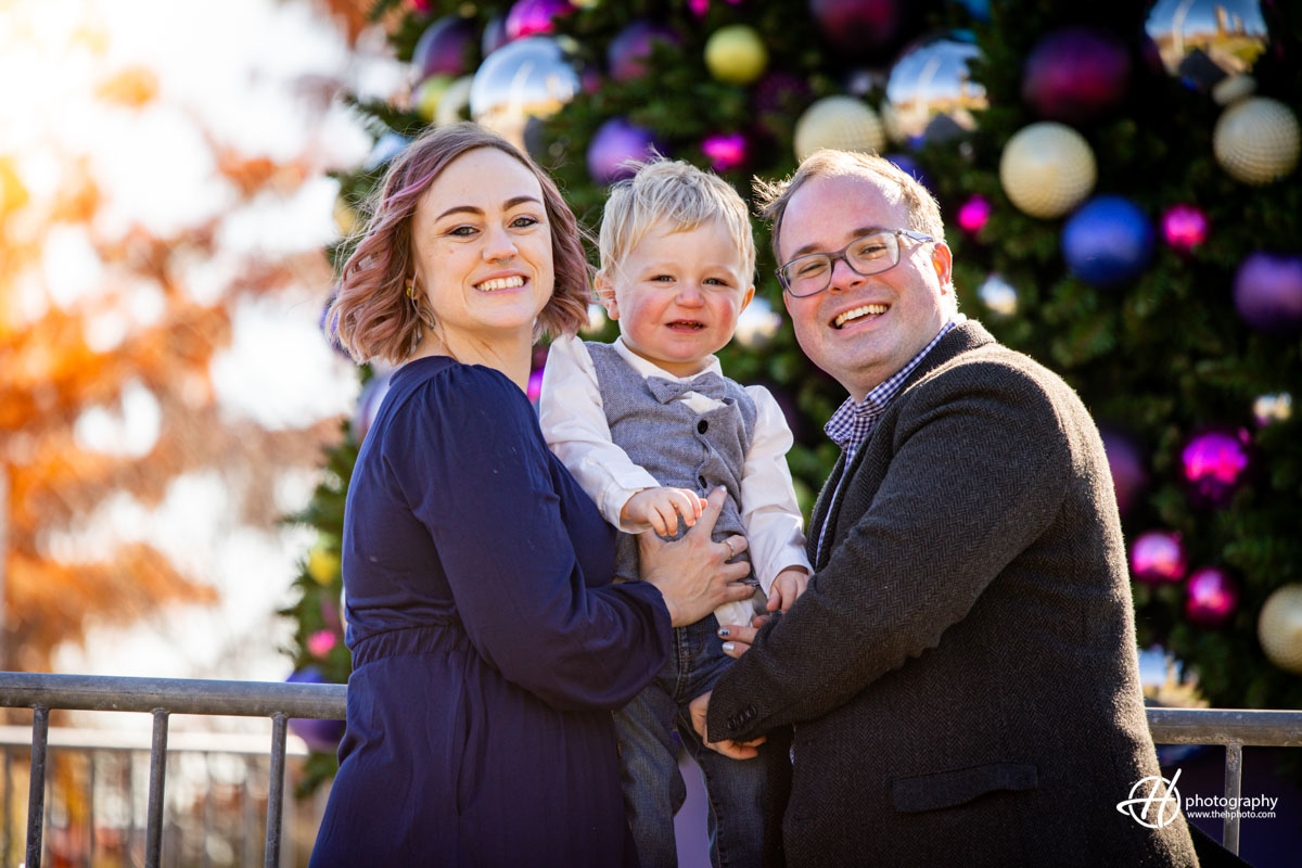 Magical holiday moment: Sara and Todd embrace their baby boy in front of a beautifully adorned Christmas tree at Navy Pier. The festive lights twinkle, casting a warm glow on the family, capturing a heartwarming scene filled with love, joy, and the spirit of the season.