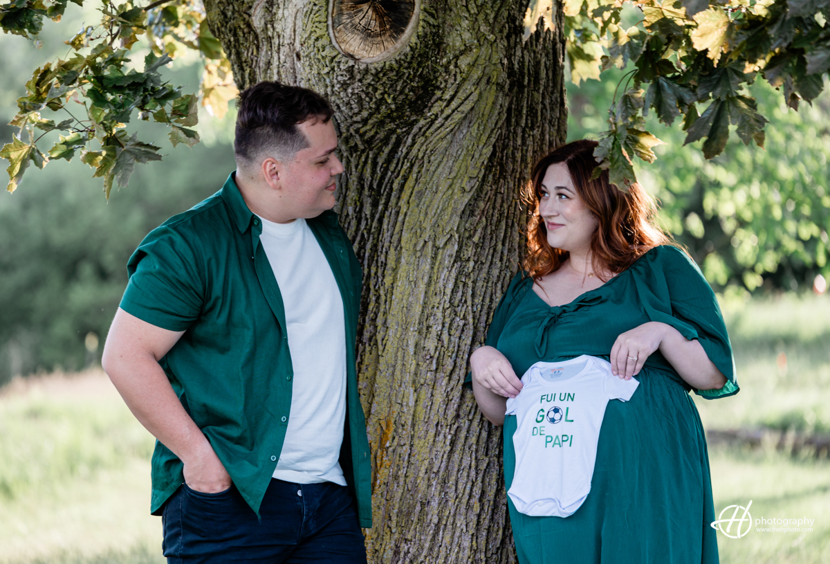 Bridget and Alejandro are seated together, holding a small T-shirt between them. The T-shirt has the phrase "Fun un GOL de Papi" written in green. Both are smiling warmly, gazing at the shirt with a sense of joy and anticipation. Bridget's long dark green dress and Alejandro's matching outfit create a cohesive look. The image captures a tender moment, symbolizing their excitement and dreams for their upcoming baby.