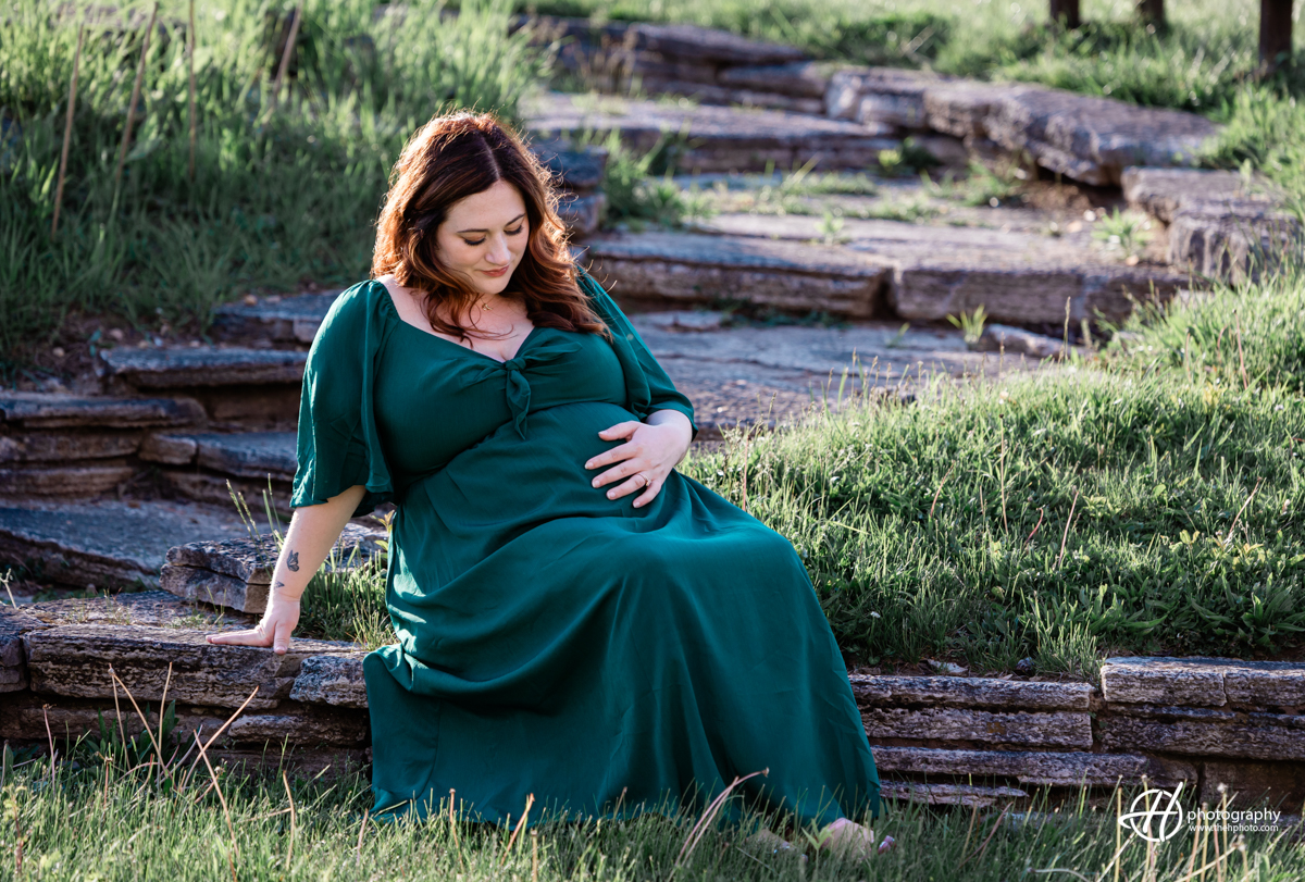 Bridget is sitting on a low stone wall, gently cradling her pregnant belly with both hands. She is wearing a long dark green dress and gazes down at her belly with a peaceful and loving expression. The background features lush greenery, highlighting the serene and natural setting of the maternity photo shoot. The image beautifully captures Bridget's anticipation and the tender connection she shares with her unborn baby.