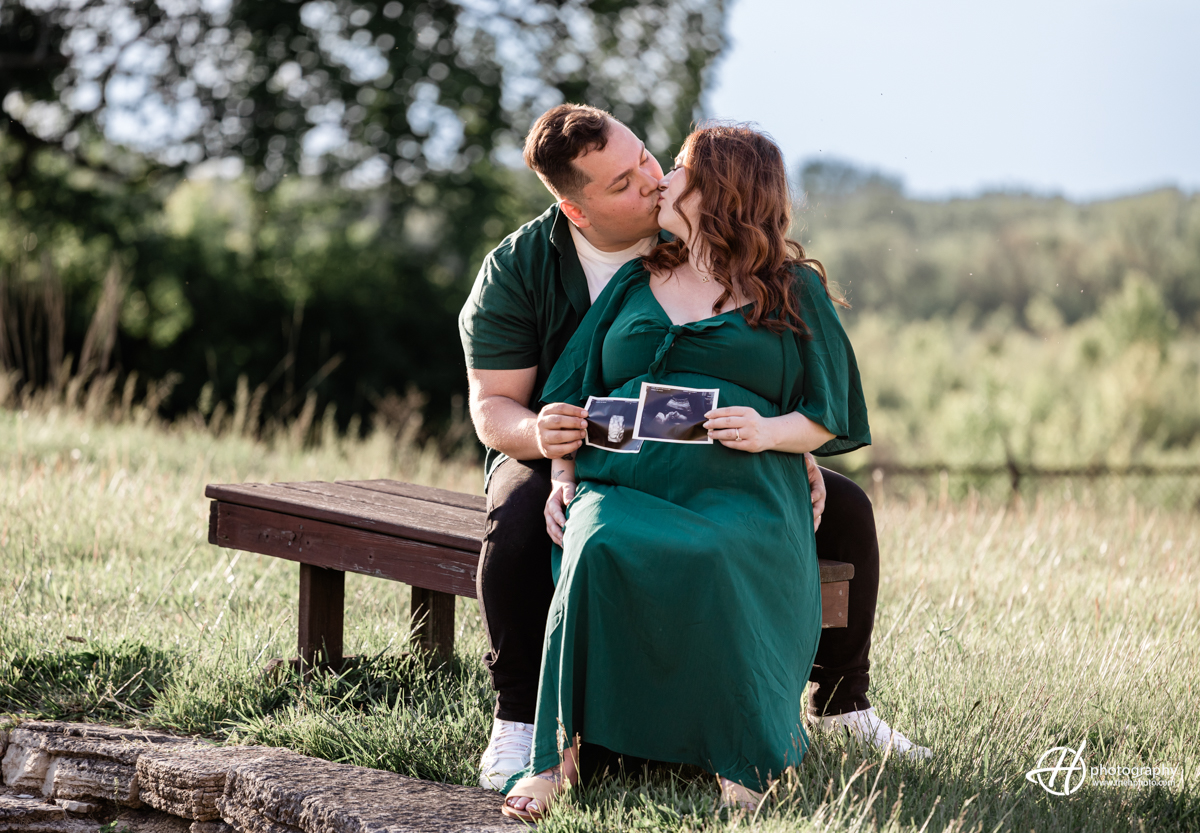 Bridget and Alejandro are sitting closely together on a wooden bench, sharing a tender moment as they look at ultrasound pictures of their baby. Bridget, dressed in a long dark green dress, holds the photos while Alejandro, in a dark green shirt and black pants, leans in to see them. Both have joyful expressions, and the serene park setting with trees and greenery in the background adds to the peaceful and intimate atmosphere of the image.