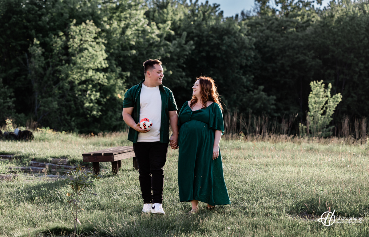 Bridget and Alejandro are walking side by side, engaged in a lively conversation. Bridget is wearing a long dark green dress, and Alejandro is dressed in a dark green shirt and black pants. Alejandro is holding a mini soccer ball in one hand. Both are smiling and their body language reflects a warm and playful connection. The background shows a scenic outdoor path with lush greenery, capturing the joy and anticipation of their journey into parenthood.