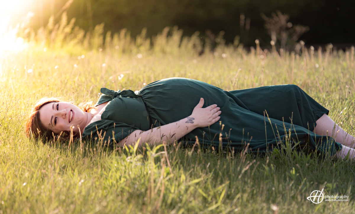 Bridget is lying on her back in the grass, surrounded by the warm rays of the setting sun. She is wearing a long dark green dress and has one hand resting on her pregnant belly. The soft sunlight creates a halo effect around her, highlighting her peaceful expression and the serene moment. The lush grass and the golden light add a magical touch to the image, capturing the beauty and tranquility of this special time in her pregnancy.