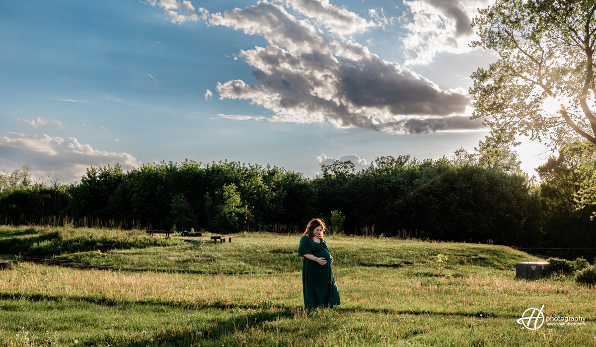 Bridget stands in a nature-filled background during sunset in the Algonquin area, admiring her pregnant belly with a gentle smile. She is wearing a long dark green dress, and the golden sunset light casts a warm glow around her. The natural scenery, with trees and soft grass, enhances the serene and intimate moment as she embraces the beauty of her pregnancy.